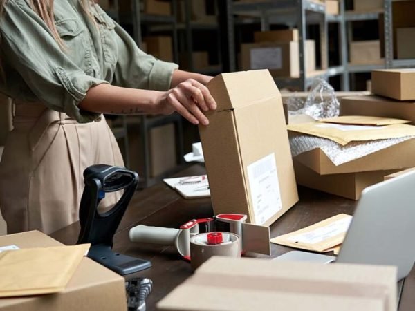 Female small business owner, entrepreneur, shipment delivery dropshipping service worker packing ecommerce order package in shipping cardboard box standing at table in warehouse. Close up view